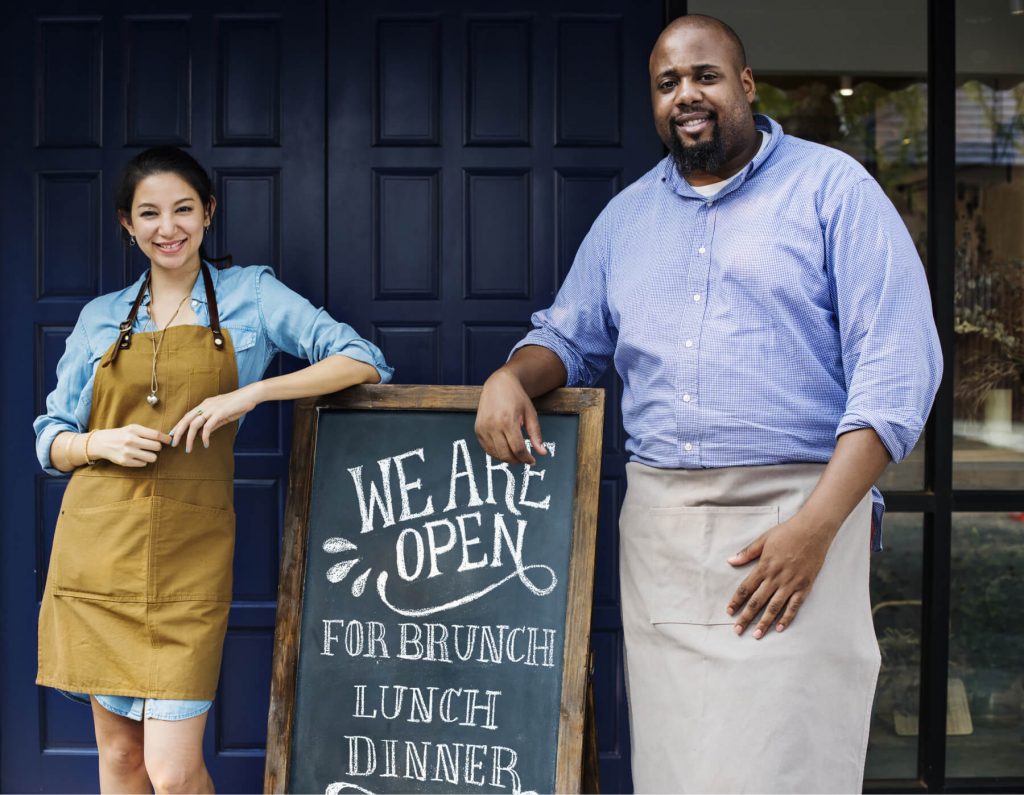Restaurant owners standing in fron of a We Are Open sign
